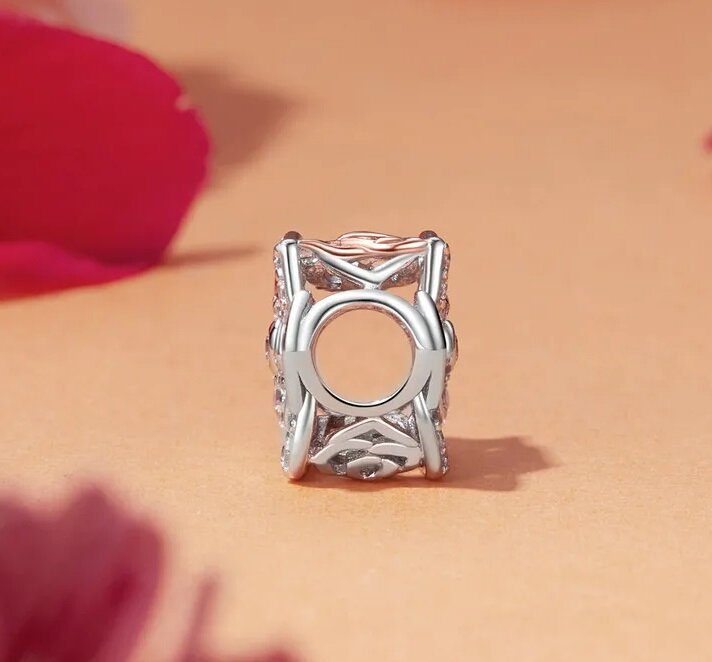 Na obrazie widoczna jest zawieszka charms w kolorze srebrnym z ażurowym wzorem, umieszczona na jasnym tle. Wokół znajdują się rozmyte elementy w kolorze różowym.