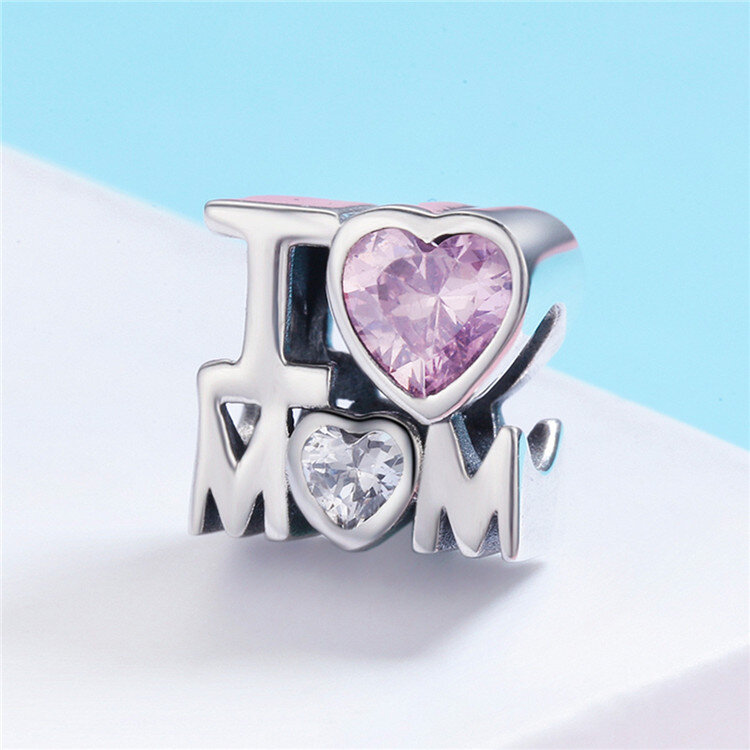 Zawieszka charms z napisem \'I LOVE MOM\', gdzie litera \'O\' jest zastąpiona różowym sercem, a \'O\' w \'MOM\' białym sercem. Tło jest jasnoniebieskie, a zawieszka ma metaliczny połysk.