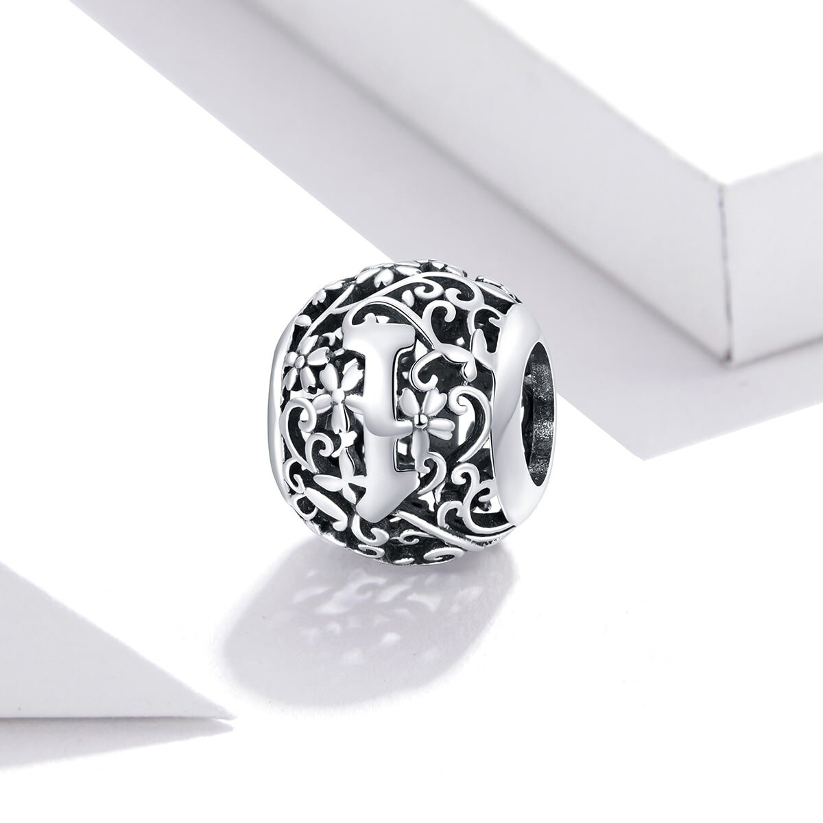 Srebrna zawieszka charms z literą \'I\' otoczona ozdobnym, ażurowym wzorem z motywem kwiatowym. Produkt jest sfotografowany na jasnym tle z geometrycznymi kształtami.
