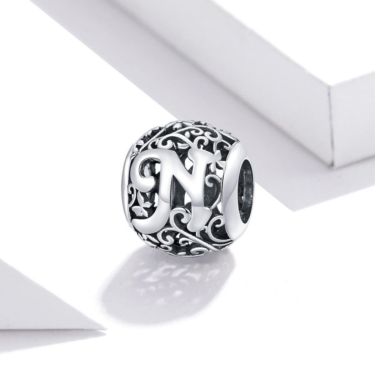 Srebrna zawieszka charms z literą \'N\' ozdobiona misternymi wzorami. W tle widoczne są białe geometryczne kształty.