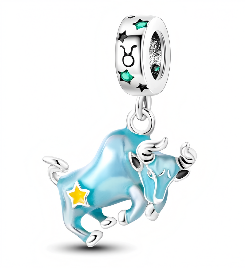 Zawieszka charms przedstawiająca niebieskiego byka z żółtą gwiazdą na boku. Na pierścieniu widoczny jest symbol znaku zodiaku Byk oraz czarne gwiazdki i zielone kamienie.