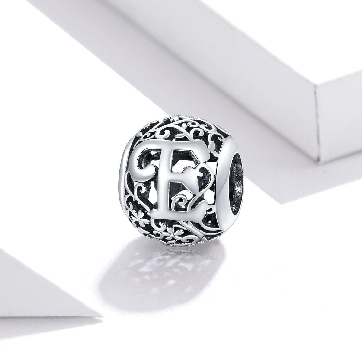 Srebrna zawieszka charms z literą \'E\' otoczona ozdobnym, ażurowym wzorem. W tle widoczne są białe, geometryczne kształty.