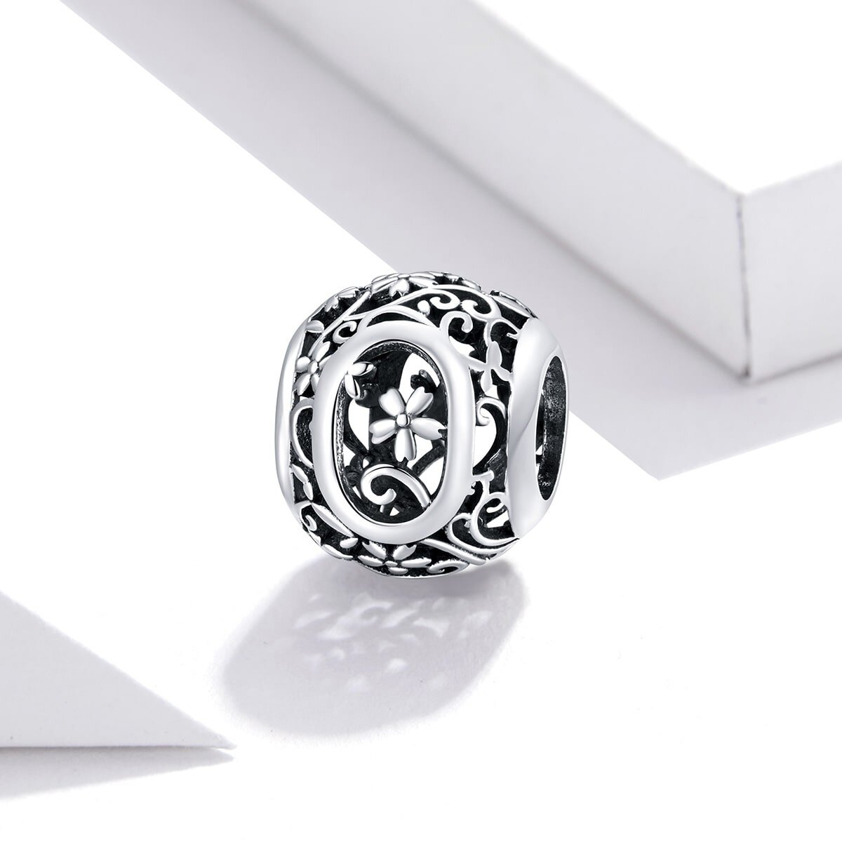 Srebrna zawieszka charms z literą \'O\' ozdobiona kwiatowym wzorem. Wykonana z metalu o błyszczącej powierzchni, prezentowana na jasnym tle.