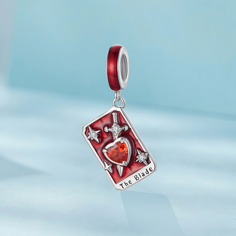 Zawieszka charms w kształcie karty tarota z czerwonym kamieniem w kształcie serca i napisem \'The Blade\'. Elementy dekoracyjne obejmują srebrne gwiazdy i miecz na czerwonym tle.