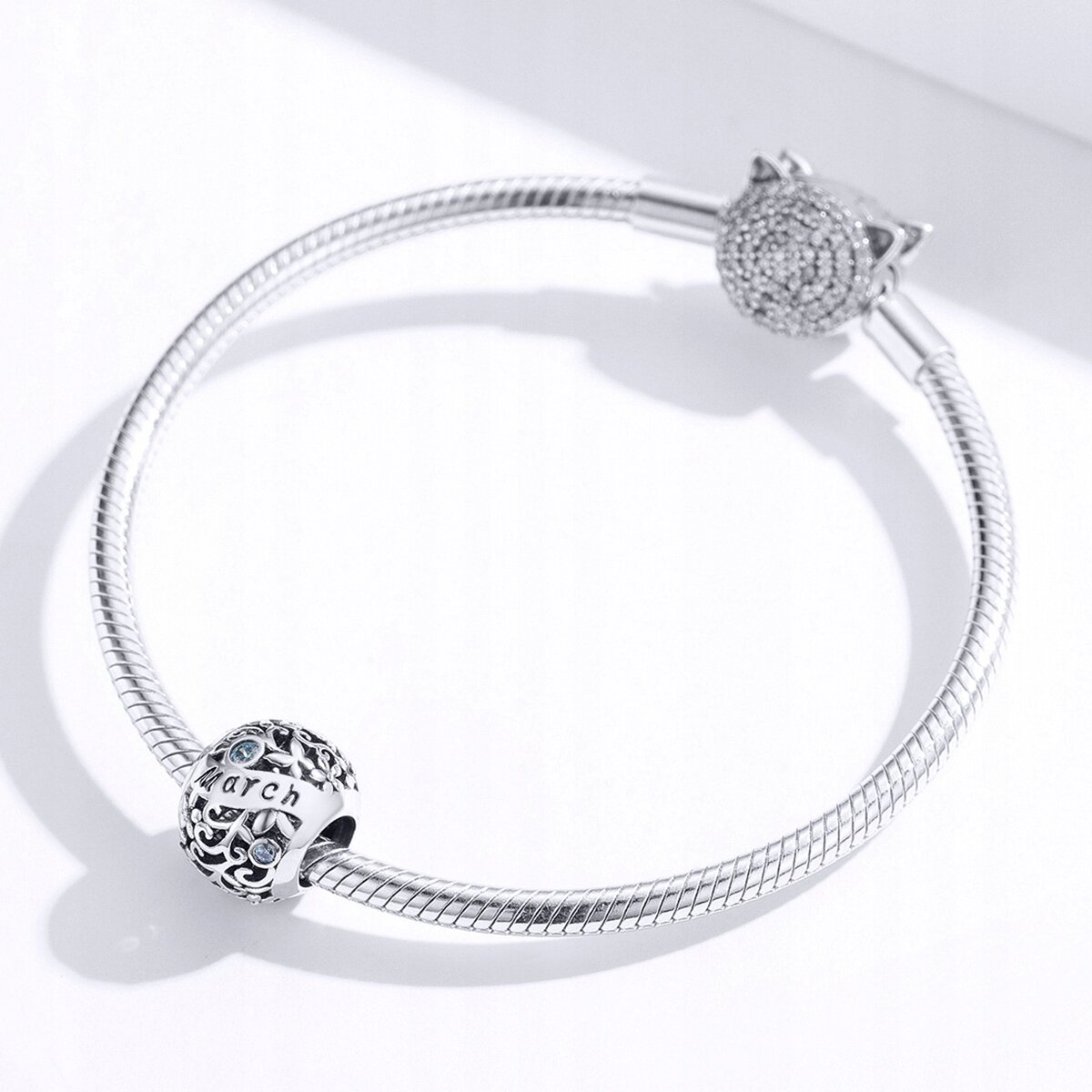 Srebrna bransoletka z charmsami, w tym jeden z napisem \'March\' i niebieskimi kamieniami. Zapięcie ozdobione jest wzorem przypominającym głowę kota.