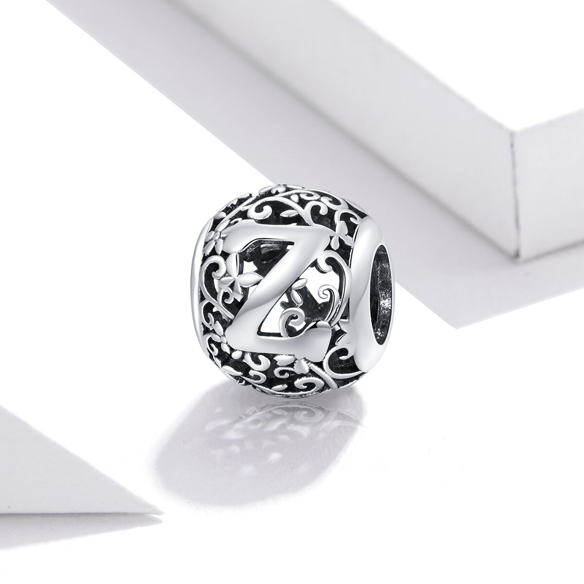 Srebrna zawieszka charms z literą \'Z\' otoczona ozdobnymi wzorami roślinnymi. Zawieszka ma ażurową strukturę i jest sfotografowana na białym tle.