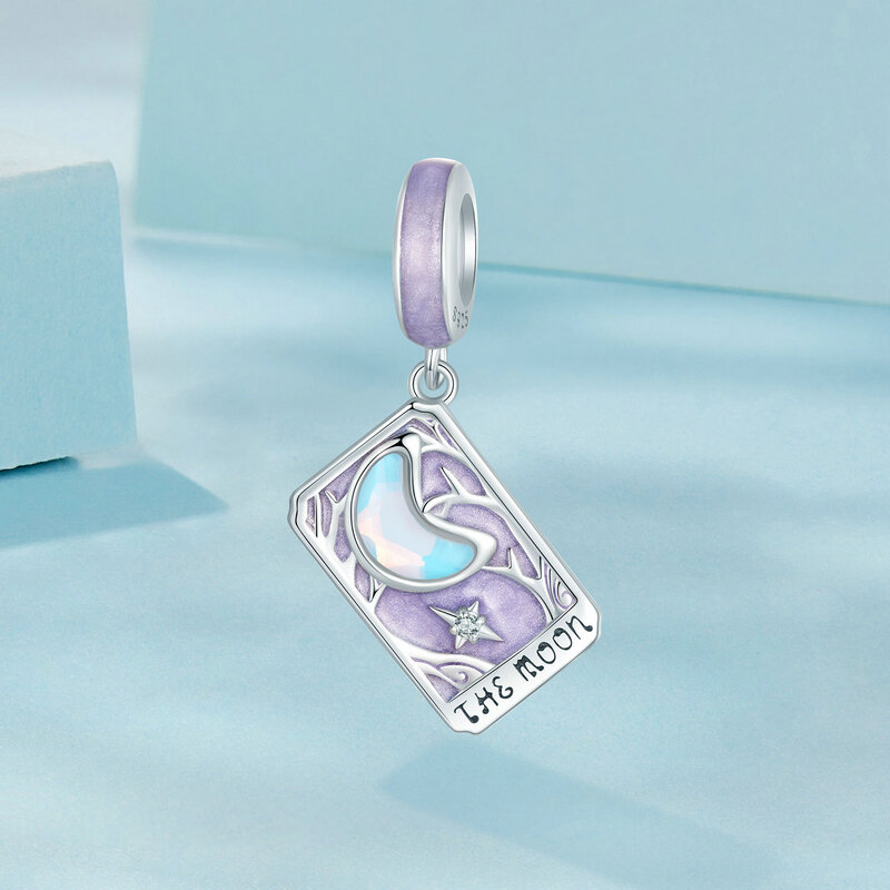 Zawieszka charms w kształcie karty tarota z napisem \'THE MOON\', ozdobiona motywem księżyca i gwiazdy. Wykonana z połyskującego srebra z fioletowymi akcentami.