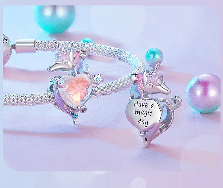 Na obrazie widoczne są srebrne zawieszki w kształcie jednorożca z różowym sercem oraz napisem \'Have a magic day\'. W tle znajdują się kolorowe, perłowe kule.