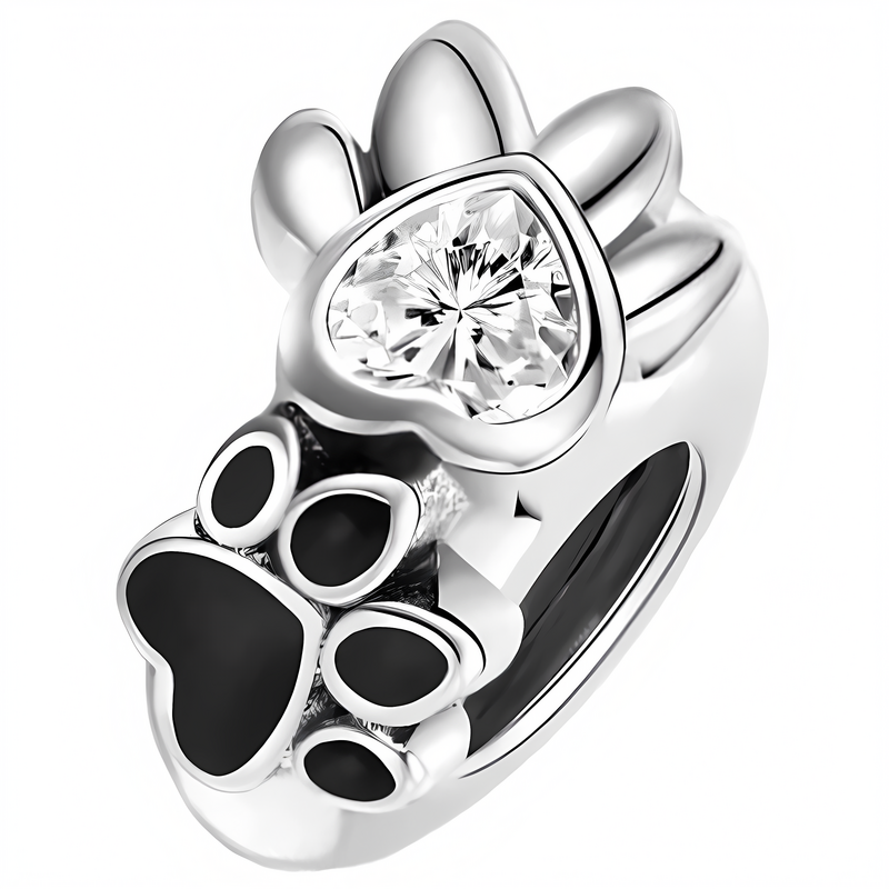 Srebrny charms w kształcie łapki zwierzęcej z czarnymi elementami i błyszczącym kamieniem w kształcie serca. Charms ma gładką, metaliczną powierzchnię.