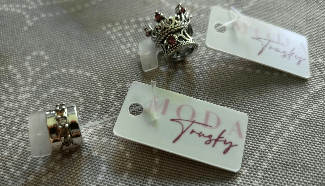 Na obrazie widoczne są trzy zawieszki w kształcie korony z czerwonymi kamieniami, każda z etykietą z napisem \'MODA Trusky\'. Zawieszki leżą na teksturowanej powierzchni.