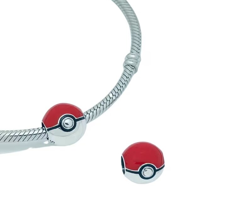 Na obrazie widoczna jest srebrna bransoletka z zawieszką w kształcie Pokeballa, obok której leży dodatkowa zawieszka o tym samym wzorze. Zawieszki mają czerwono-białe kolory z czarnym paskiem i srebrnym środkiem.