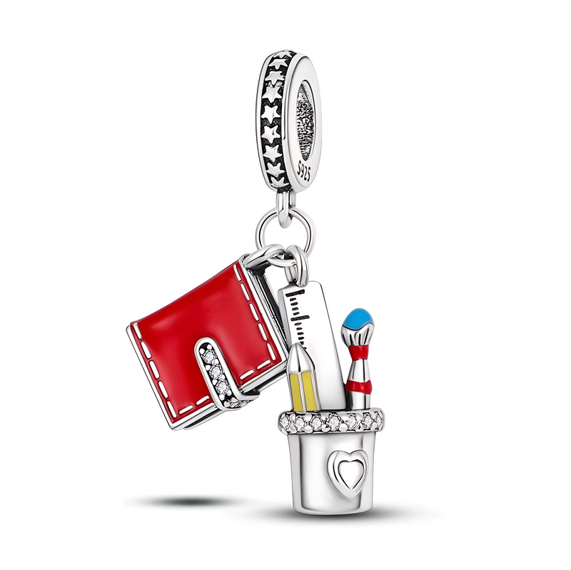 Zawieszka charms przedstawia czerwony zeszyt, linijkę, ołówek i pędzel w srebrnym kubku z sercem. Na obręczy widoczne są gwiazdki i napis \'S925\'.