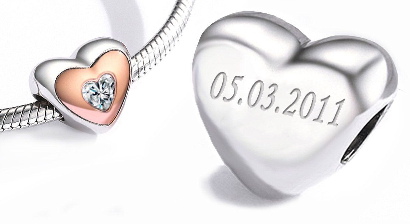 Na obrazie widoczna jest zawieszka w kształcie serca z grawerem \'05.03.2011\'. Zawieszka jest wykonana z metalu i ma ozdobny element w kształcie serca z przezroczystym kamieniem.