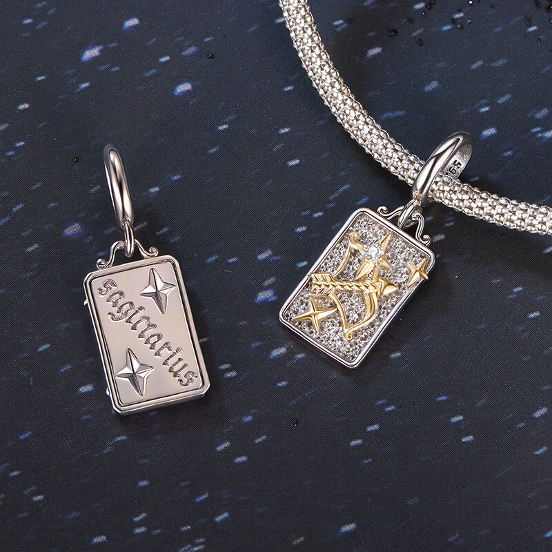 Na obrazie widoczne są dwie zawieszki charms w kształcie prostokąta, jedna z napisem \'sagittarius\' i gwiazdami, druga z motywem łucznika i gwiazdami. Obie zawieszki są wykonane z metalu, jedna z elementami w kolorze złotym, umieszczone na tle przypominającym nocne niebo.