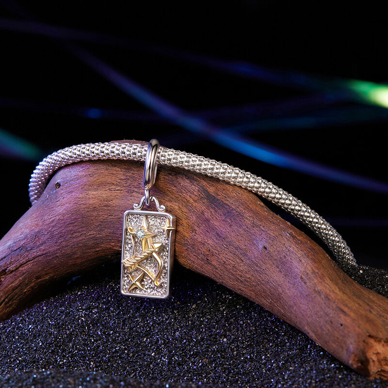 Na zdjęciu widoczna jest srebrna zawieszka charms z motywem znaku zodiaku Strzelec, umieszczona na plecionym srebrnym łańcuszku. Tło stanowi ciemne, błyszczące podłoże oraz fragment drewna.