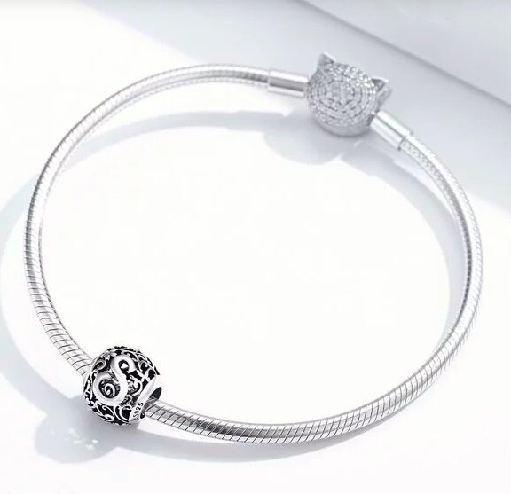 Na zdjęciu widoczna jest srebrna bransoletka z okrągłym charms z literą \'S\' o ozdobnym wzorze. Zapięcie bransoletki ma kształt głowy kota z błyszczącymi elementami.