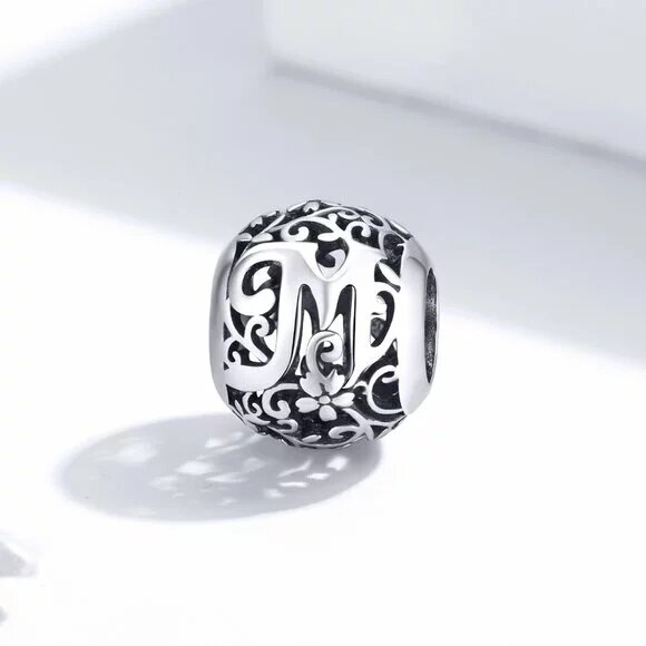 Srebrny charms z literą \'M\' otoczony ozdobnym, ażurowym wzorem. Charms ma okrągły kształt i jest wykonany z metalu o błyszczącej powierzchni.