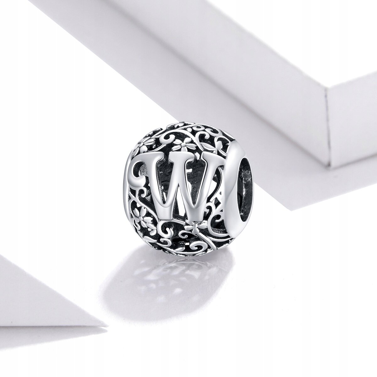 Srebrna zawieszka charms w kształcie kuli z literą \'W\' na tle ozdobnych wzorów. Wykonana z metalu o ciemnym tle i jasnych, wypukłych elementach dekoracyjnych.