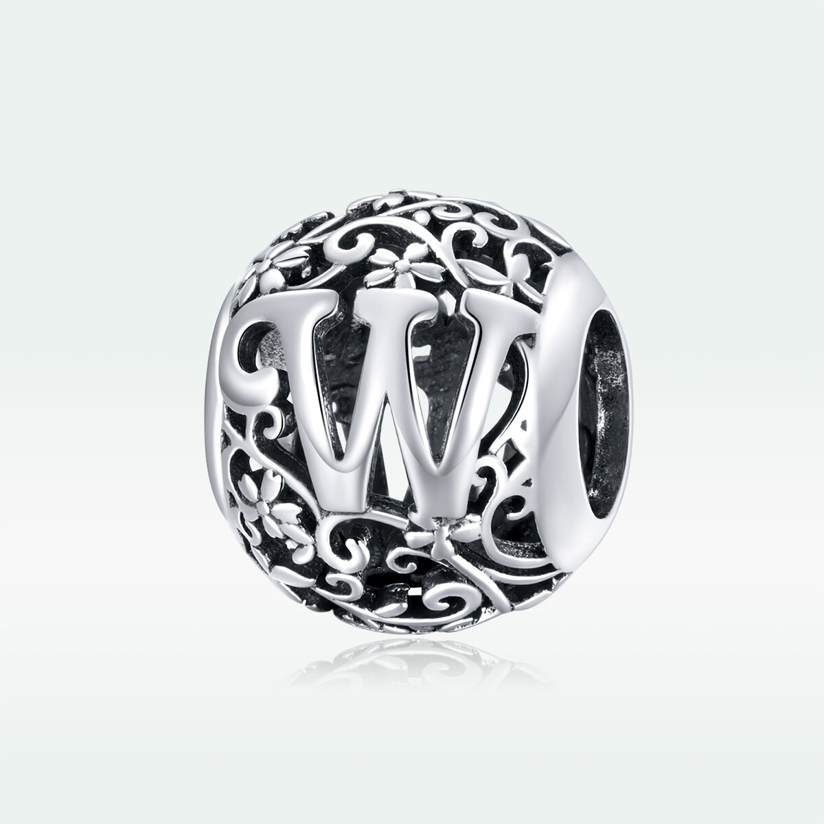 Srebrna zawieszka charms z literą \'W\' otoczona ozdobnym, ażurowym wzorem z motywem roślinnym. Wykonana z metalu o wysokim połysku.