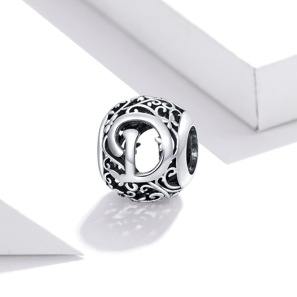 Srebrna zawieszka charms z literą \'D\' na ażurowym tle z ozdobnymi wzorami. Produkt jest sfotografowany na białym tle.