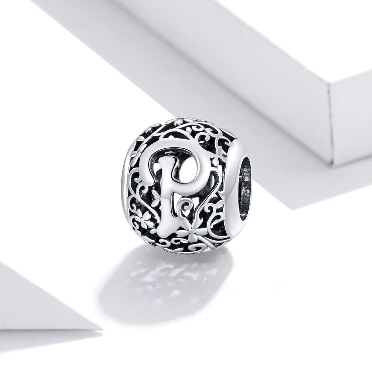 Srebrna zawieszka charms z literą \'P\' otoczona ozdobnym, ażurowym wzorem z motywem roślinnym. Zawieszka ma kształt kulisty i jest wykonana z metalu o błyszczącej powierzchni.