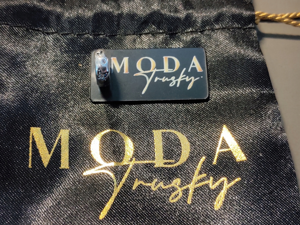 Na czarnym tle znajduje się srebrny charms z motywem motylków, umieszczony na kartoniku z napisem \'MODA Trusky\'. W tle widoczny jest woreczek z tym samym napisem w kolorze złotym.