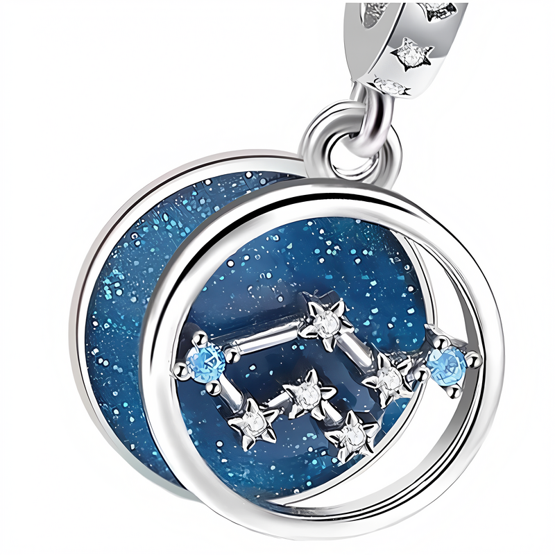 Zawieszka charms z motywem znaku zodiaku Bliźnięta, ozdobiona srebrnymi gwiazdkami i niebieskimi kamieniami na tle z brokatowym efektem. Elementy metalowe mają srebrny kolor.