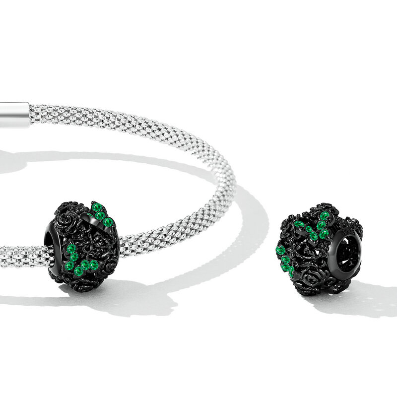 Na obrazie widoczna jest srebrna bransoletka z ażurowym charms w kształcie motyli, ozdobionym zielonymi kamieniami. Charms jest wykonany z czarnego materiału z detalami przypominającymi kwiaty.