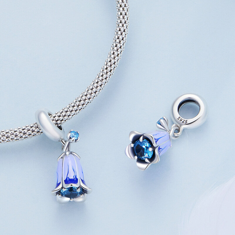 Na jasnym tle widoczny jest srebrny charms w kształcie kwiatu z niebieskim kamieniem, przymocowany do plecionego łańcuszka. Obok leży podobny charms z wygrawerowanym napisem \'S925\'.