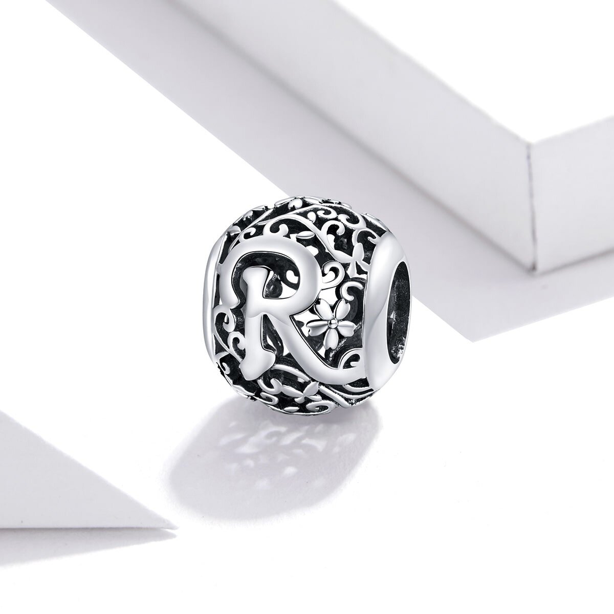 Srebrny charms z literą \'R\' na ażurowym tle z kwiatowym wzorem. Charms ma okrągły kształt i jest wykonany z metalu o błyszczącej powierzchni.
