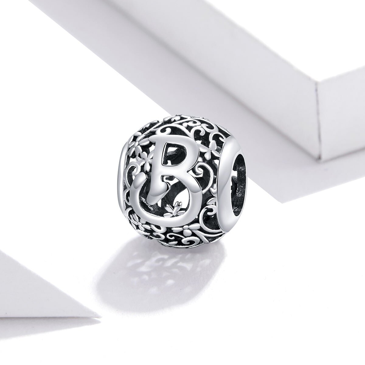 Srebrna zawieszka charms z literą \'B\' otoczona ozdobnym wzorem z motywem roślinnym. Zawieszka ma ażurową strukturę i błyszczącą powierzchnię.