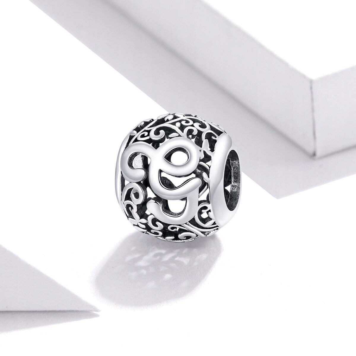 Srebrna zawieszka charms z literą \'G\' na ażurowym tle z ozdobnymi wzorami. Produkt wykonany z metalu o ciemnym i jasnym wykończeniu.