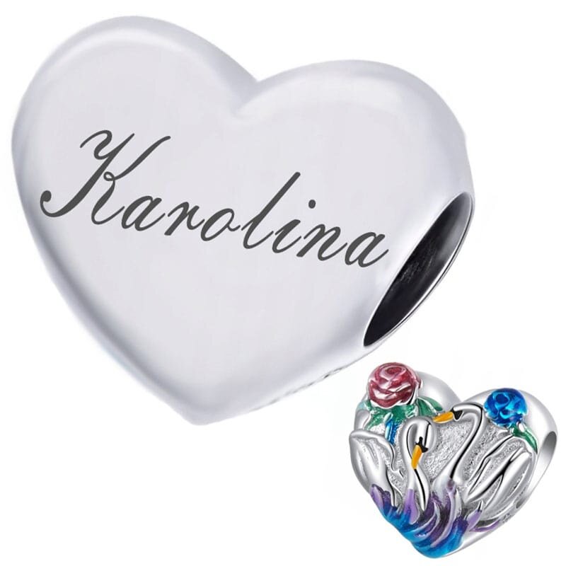 Na obrazie widoczna jest srebrna zawieszka w kształcie serca z grawerem \'Karolina\'. Obok znajduje się ozdobna zawieszka z motywem łabędzi i kolorowych kwiatów.