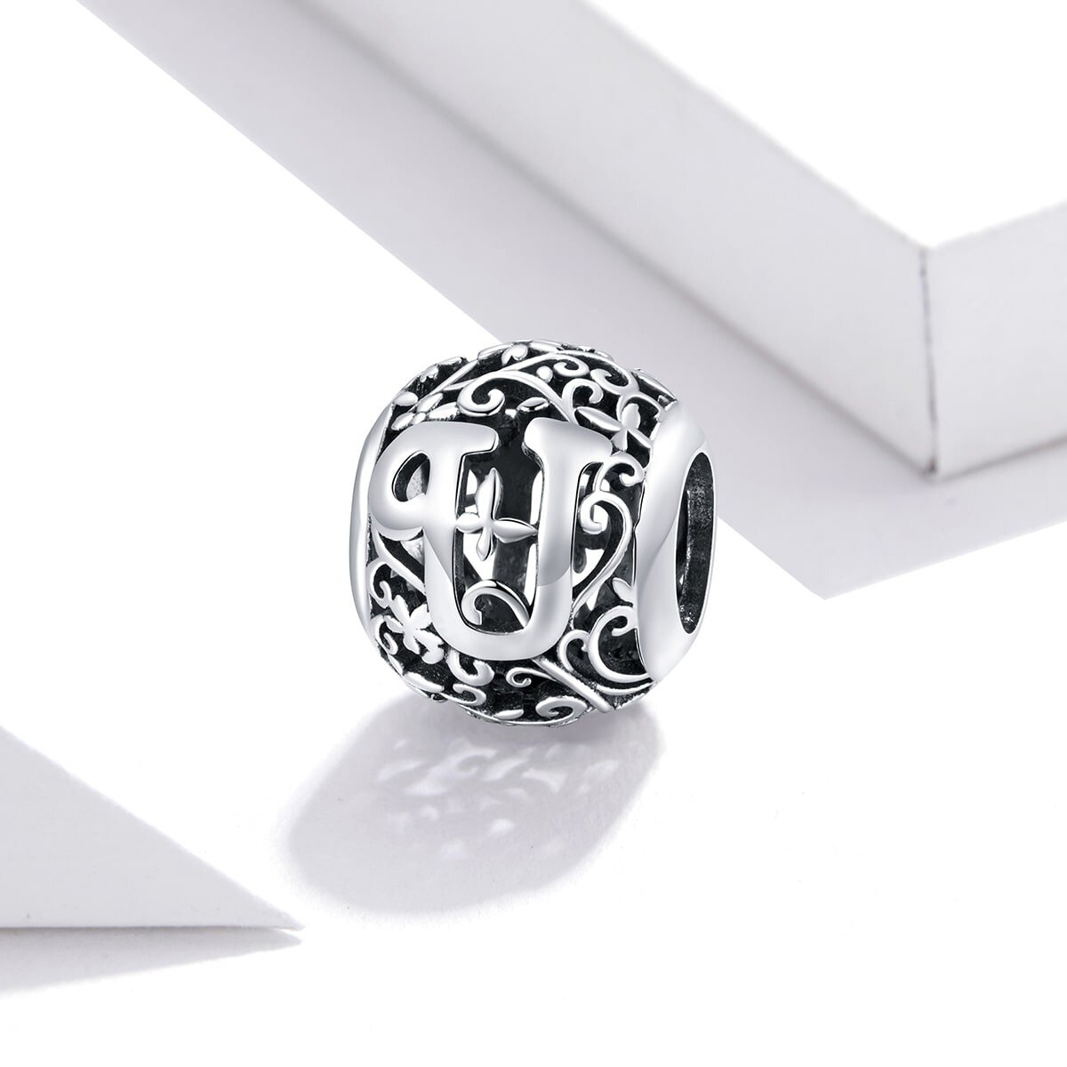 Srebrna zawieszka charms w kształcie kuli z literą \'U\' na ozdobnym tle z motywem roślinnym. Wykonana z metalu o ciemnym wykończeniu, z widocznymi detalami dekoracyjnymi.