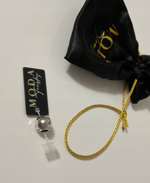 Na białym tle znajduje się srebrny koralik z czerwonymi elementami, przymocowany do czarnej metki z napisem \'MODA Jewelry\'. Obok leży czarny woreczek z żółtym sznurkiem i złotym napisem.