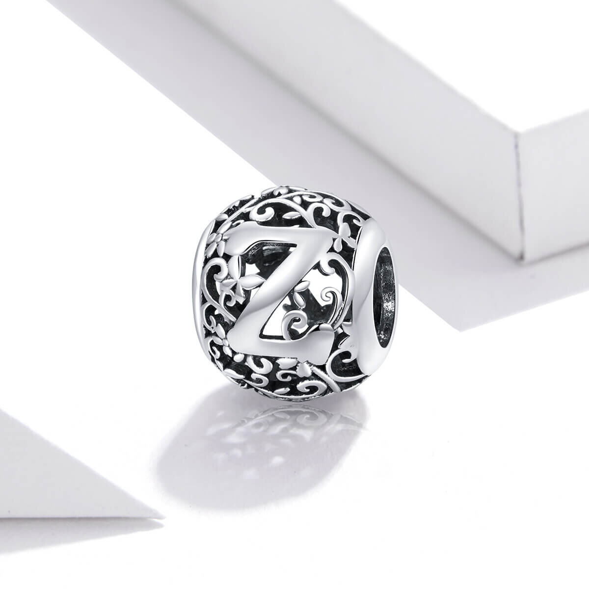 Srebrna zawieszka charms z literą \'Z\' ozdobiona delikatnymi wzorami roślinnymi. W tle widoczne są białe geometryczne kształty.