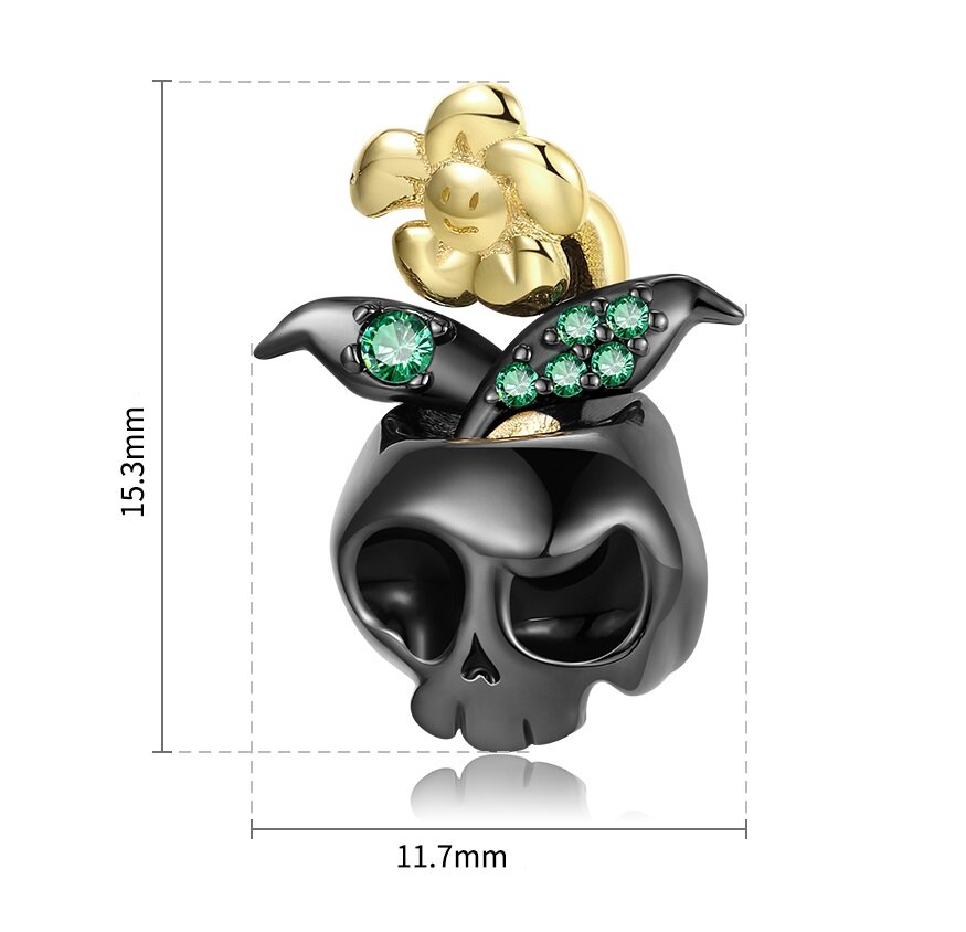 Zawieszka charms w kształcie czaszki z czarnego materiału, ozdobiona złotym kwiatem i zielonymi kamieniami. Wymiary zawieszki to 15,3 mm na 11,7 mm.