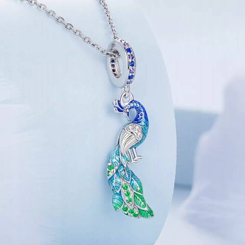 Zawieszka charms w kształcie pawia z kolorowymi piórami w odcieniach niebieskiego i zielonego, ozdobiona błyszczącymi kamieniami. Zawieszona na srebrnym łańcuszku z okrągłym elementem ozdobionym niebieskimi kamieniami.