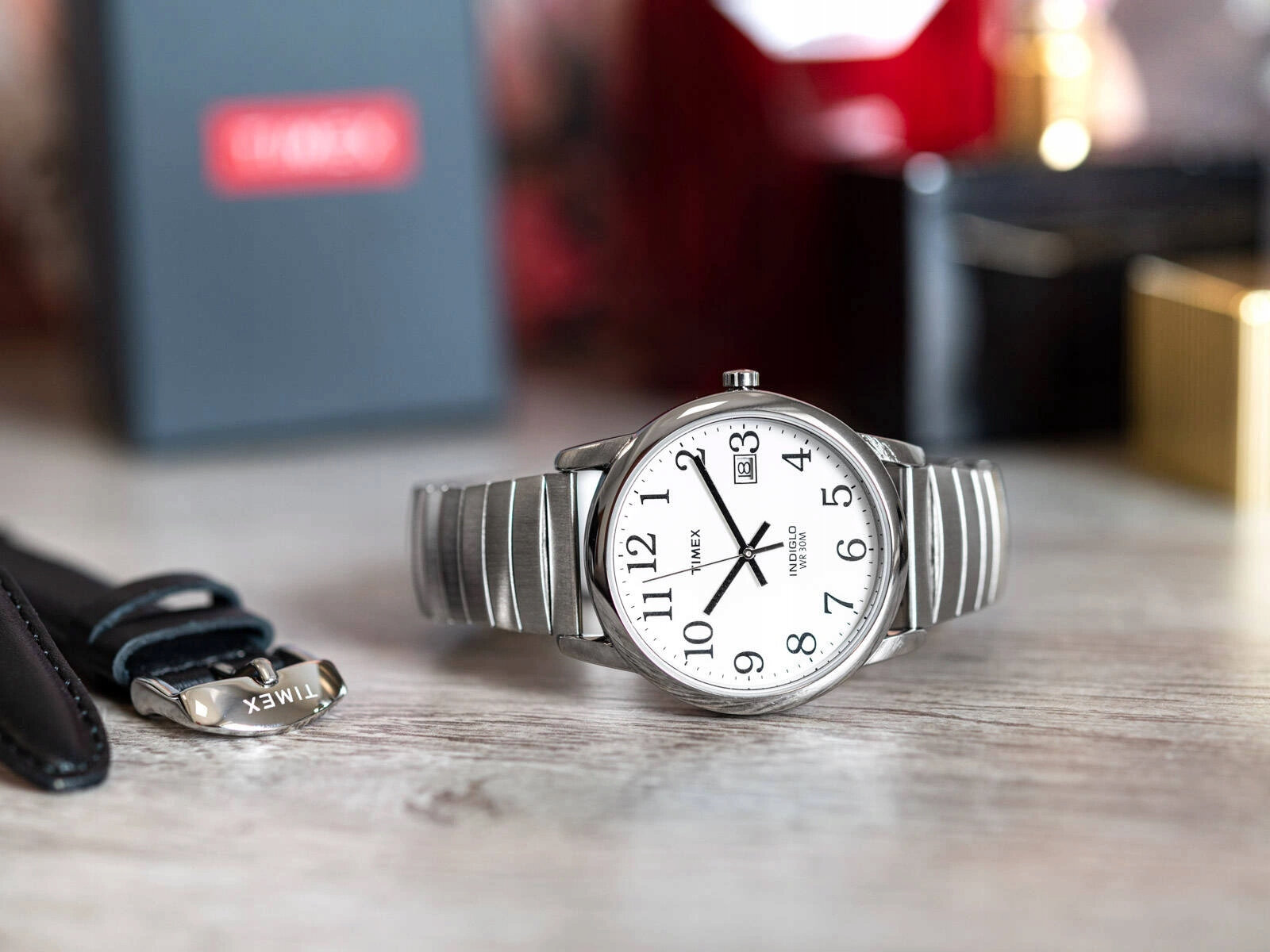 Na drewnianym blacie znajduje się zegarek z białą tarczą i metalową bransoletą, z widocznym logo \'TIMEX\'. W tle widoczny jest rozmyty czarny pudełko z czerwonym napisem \'TIMEX\'.