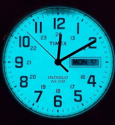 Zegarek z podświetlaną tarczą w kolorze niebieskim, z dużymi cyframi arabskimi i wskazówkami. Widoczny jest napis \'TIMEX\', \'INDIGLO\', \'WR 30M\' oraz okienko z dniem tygodnia \'MON\'.