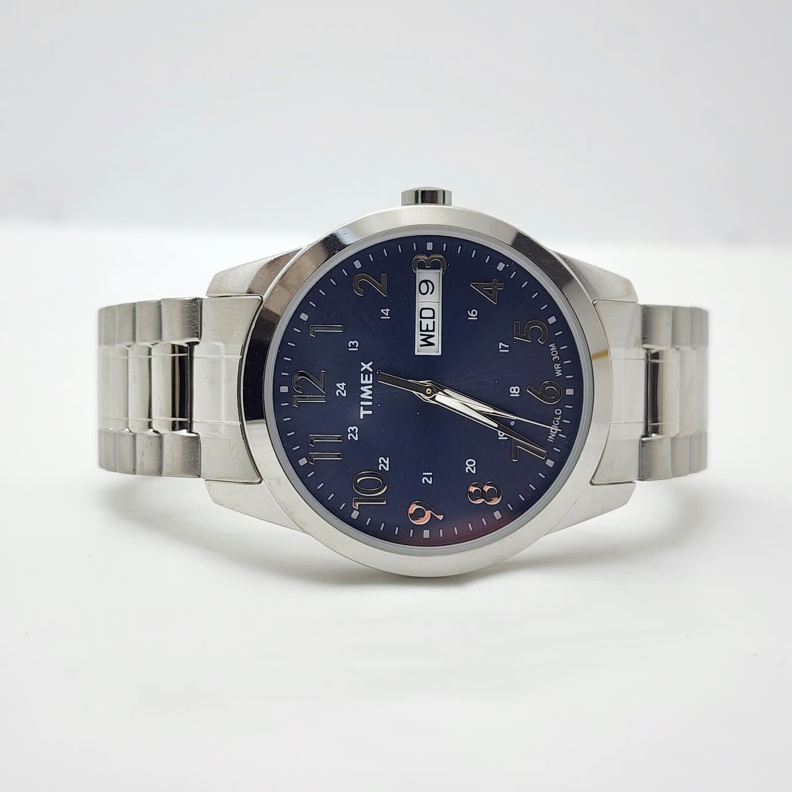 Zegarek z metalową bransoletą i ciemną tarczą, na której widoczne są srebrne wskazówki oraz cyfry. Na tarczy znajduje się napis \'TIMEX\' oraz okienko z dniem tygodnia i datą.