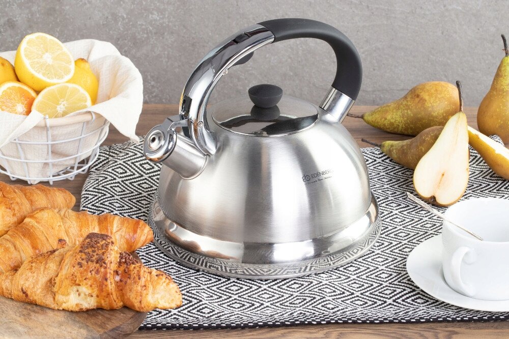 Na obrazie znajduje się stalowy czajnik z czarnym uchwytem, umieszczony na wzorzystej podkładce. W tle widoczne są croissanty, pokrojone gruszki, filiżanka oraz kosz z cytrynami i pomarańczami.