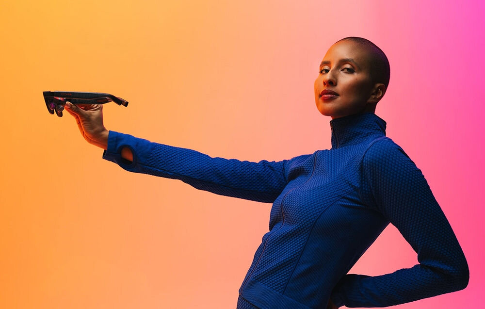 Okulary VITURE XR Luma Ultra Czarny Kobieta w granatowej bluzie trzyma okulary VR w dłoni, za nią pomarańczowo-różowe tło zestaw kamer śledzenie otoczenia kamera RGB podwójne kamery głębi obsługa gestów tryb 6DoF
