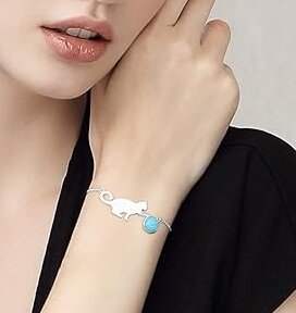 Na zdjęciu widoczna jest srebrna bransoletka z motywem kotka i niebieską kulką, noszona na nadgarstku osoby ubranej w czarną odzież. Bransoletka wykonana jest z jasnego metalu i ma delikatny, elegancki wygląd.
