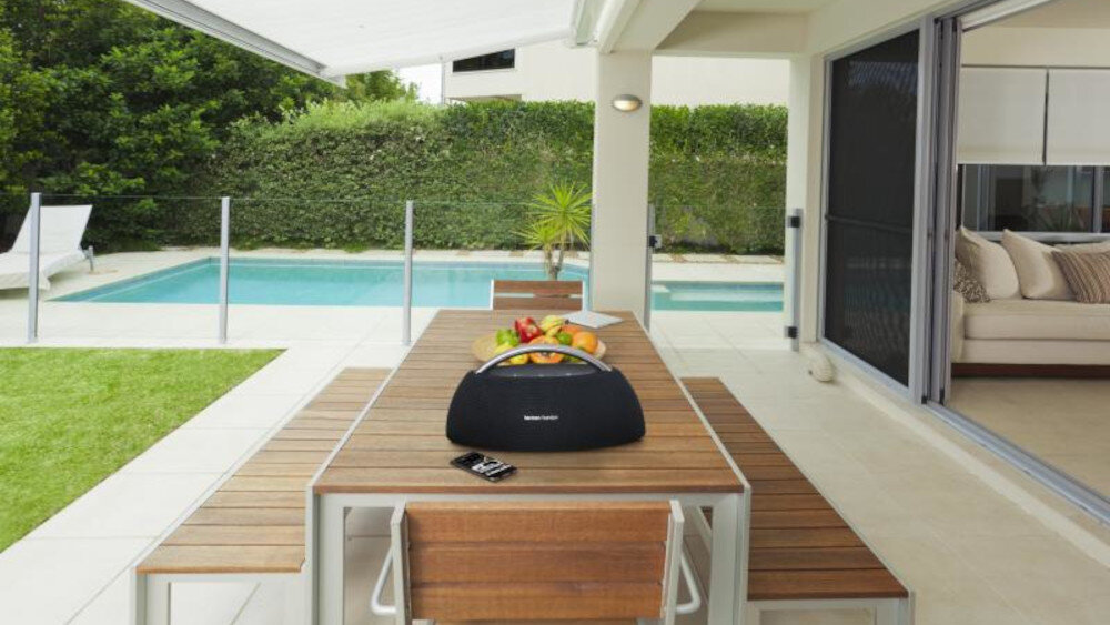 Na drewnianym stole w ogrodzie stoi czarny głośnik z logo \'Harman Kardon\'. W tle widoczny jest basen oraz część domu z otwartym salonem.