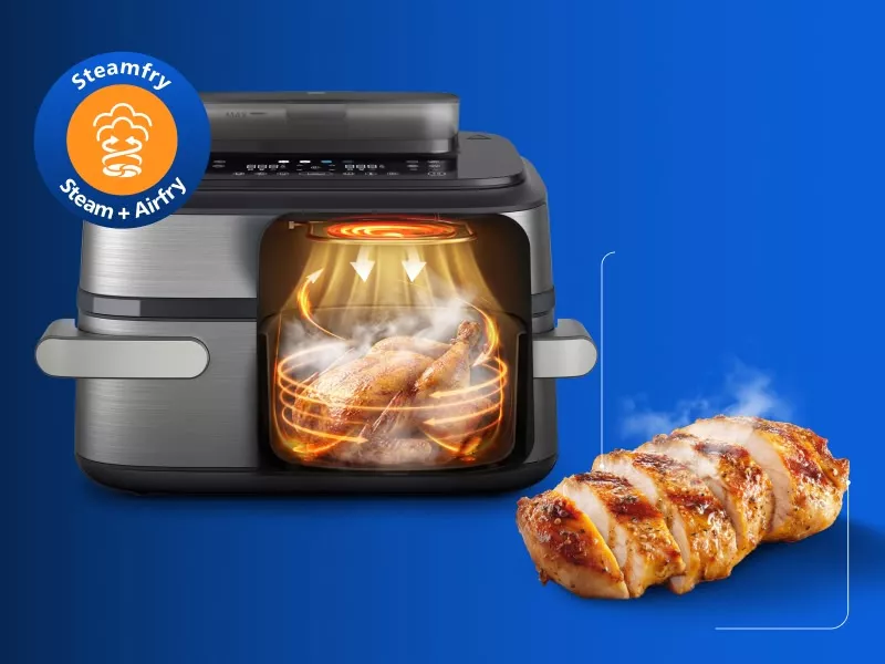 Obraz przedstawia frytkownicę beztłuszczową z funkcją Steamfry, w której widać pieczonego kurczaka. Obok urządzenia znajduje się pokrojony kawałek mięsa, a w rogu widnieje logo z napisem \'Steamfry Steam + Airfry\'.