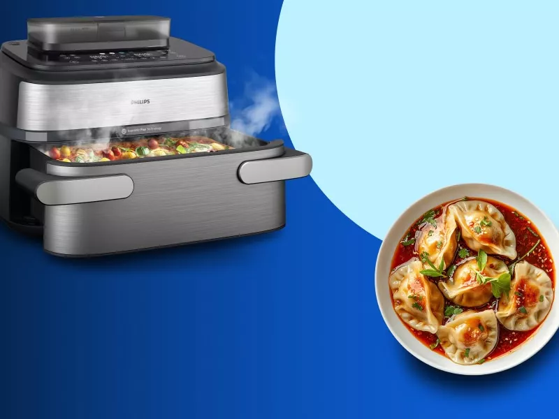 Na niebieskim tle widoczna jest frytkownica beztłuszczowa z otwartą szufladą, z której wydobywa się para. Obok znajduje się talerz z pierożkami w sosie.