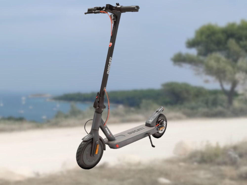 Na obrazie widoczna jest czarna hulajnoga elektryczna z napisem \'DUCATI\' na pionowej części kierownicy. W tle znajduje się rozmyty krajobraz z drzewami i wodą.