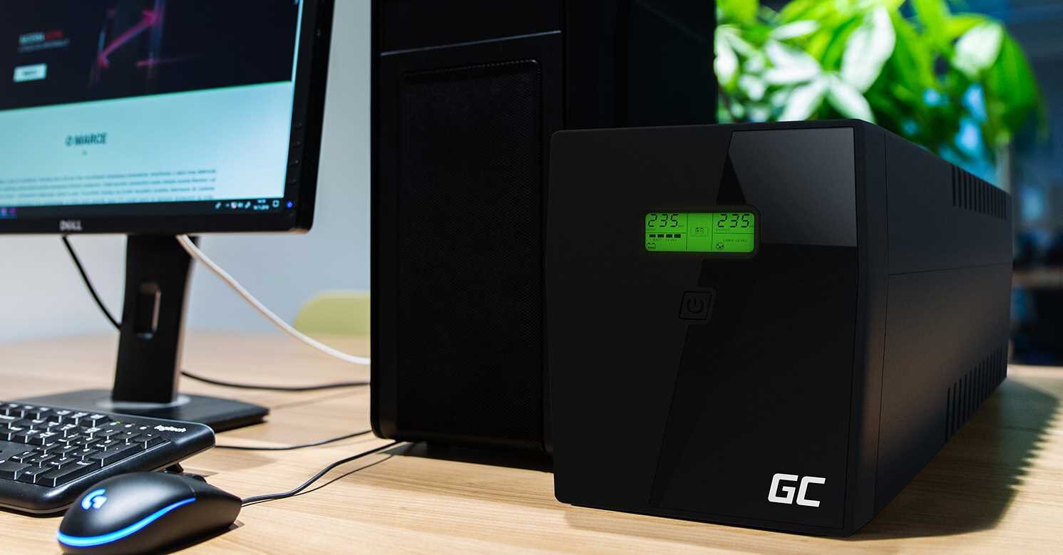 Na biurku znajduje się zasilacz UPS z wyświetlaczem LCD pokazującym wartości 235 i 235 oraz logo GC. Obok stoi monitor, klawiatura i myszka.