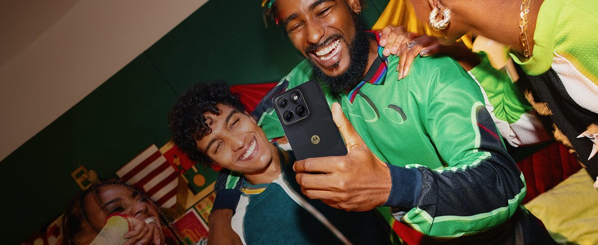 Grupa osób ubranych w kolorowe stroje trzyma smartfon z widocznym logo producenta. W tle znajdują się kolorowe flagi i dekoracje.